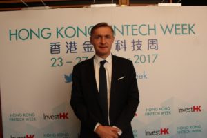 A man in a black suit and tie standing in front of a Hong Kong FinTech Week banner.