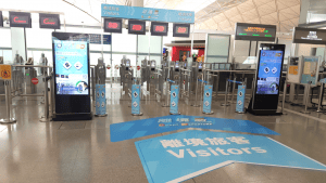 A security checkpoint with turnstiles and digital screens at an airport or transit hub.