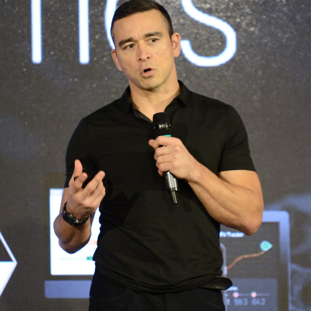 Man in black shirt speaking on stage with a microphone.