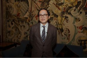 Man in glasses and suit standing against a decorative wall.