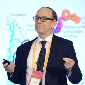 A man in a suit giving a scientific presentation with molecular diagrams in the background.