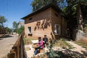 People sitting and talking outside a rustic brown house on a sunny day.