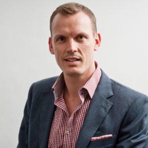 Professional man in a blazer and checkered shirt posing against a plain background.