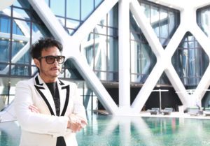 Man in stylish white suit stands by a modern pool with glass and steel architecture.