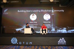 Two speakers discuss blockchain at a conference stage with a large screen behind them.