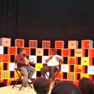 Two men in discussion on stage with a colorful cube backdrop.