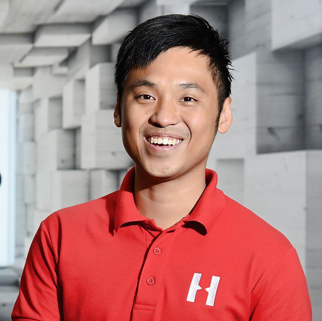 Smiling man in a red polo shirt with an H logo, standing indoors.