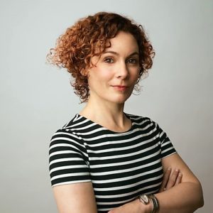 Confident woman with curly hair in a striped shirt posing with crossed arms.