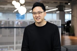 Young man wearing glasses and a black sweater, smiling indoors.