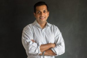 Smiling man in a light shirt poses with crossed arms against a dark background.