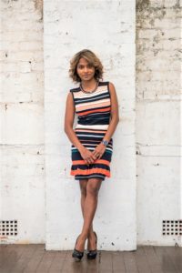 Woman in a striped sleeveless dress standing against a white brick wall.