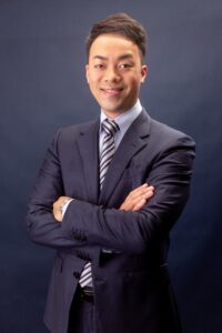 Confident young businessman in a suit with arms crossed, smiling against a solid background.
