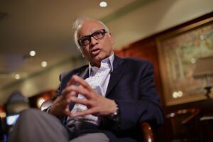An elderly man in glasses and a suit speaks thoughtfully indoors.