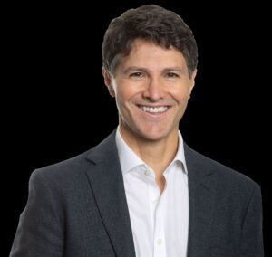 Smiling man in a dark blazer and white shirt against a black background.