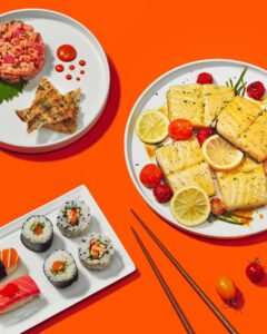 A vibrant spread of sushi, grilled fish, and garnishes on an orange table.