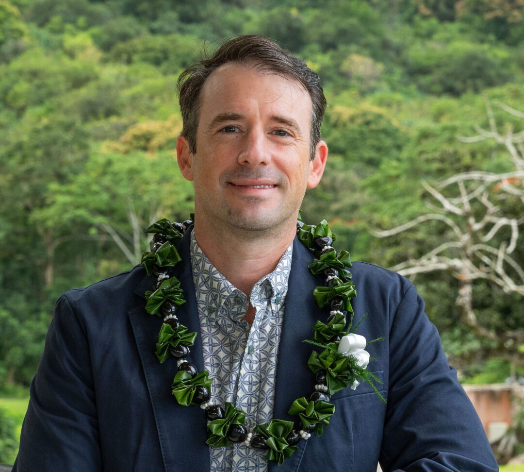 Man in a suit with a traditional Hawaiian lei, smiling outdoors.
