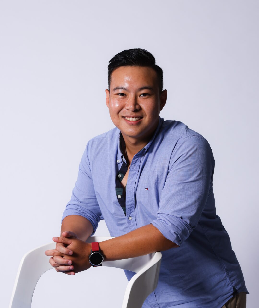 Smiling man in a blue shirt leaning on a white chair.