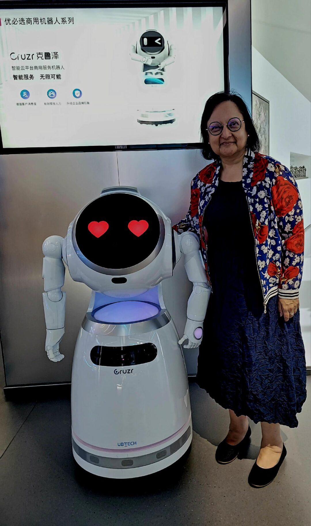 A woman stands next to a white robot with heart-shaped eyes in a tech exhibit.