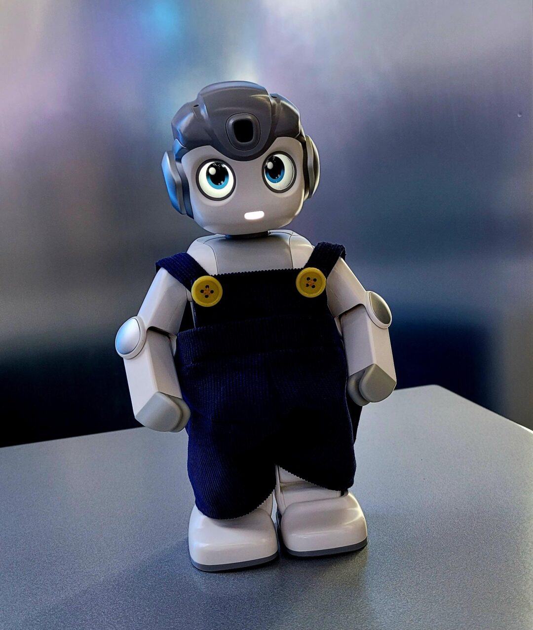 A small robot with big blue eyes wearing dark overalls stands on a table.