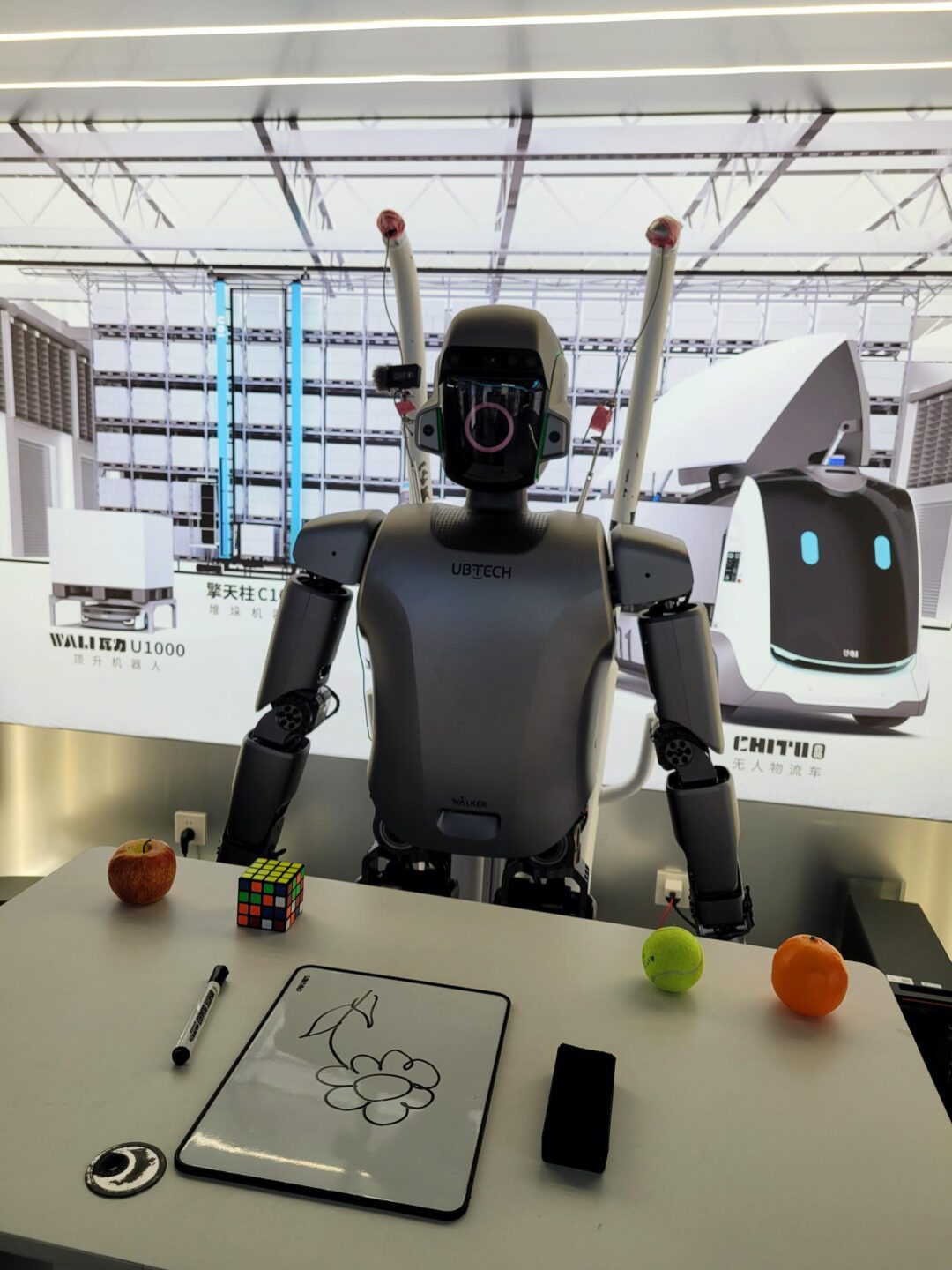 A UBTECH robot stands behind a table with various objects and a drawing board.