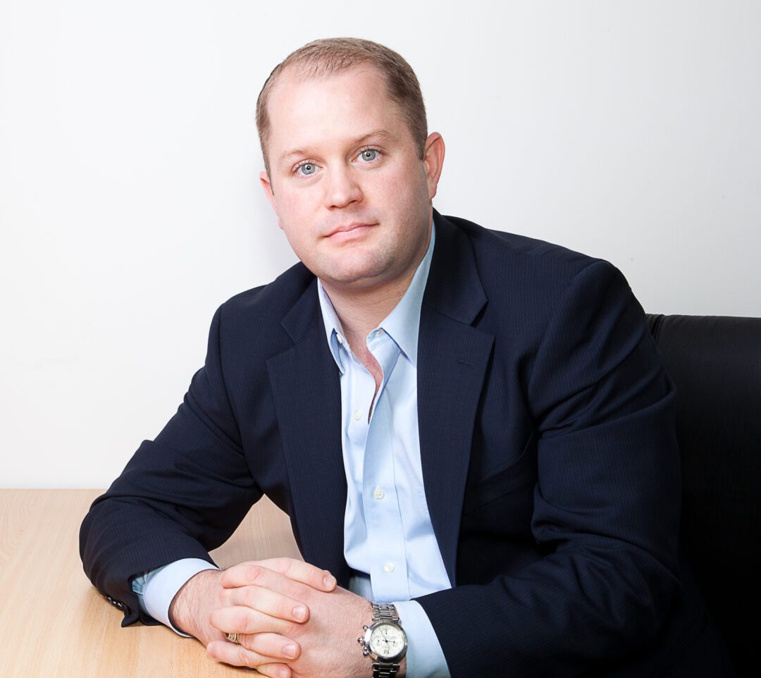 Man in a navy blazer sitting at a desk, looking at the camera.